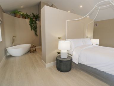 A bedroom with a white bed and two lamps next to a partition wall with a bathtub and plants behind it at The Barn in Llanfechell