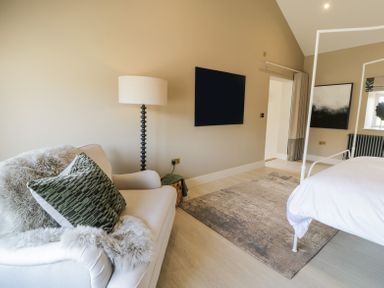A bedroom with a white armchair with cushions a floor lamp a mounted television a bed and a rug at The Barn in Llanfechell