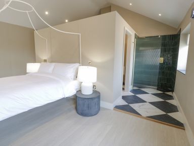 A bedroom with a white bed and lamps next to a bathroom with black and white checkered floor at The Barn in Llanfechell