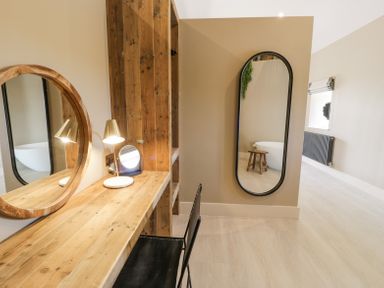 A wooden desk with a round mirror lamp and chair and a tall oval mirror on the wall in a room at The Barn in Llanfechell
