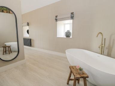 A bathroom with a white freestanding bathtub a wooden stool with a small flower in a glass a tall mirror a window with a vase on the sill and a radiator at The Barn in Llanfechell