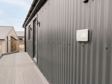 An outdoor corridor with gray decking beside a corrugated metal wall with a sign reading The Gym at Farmyard House in Llanfechell