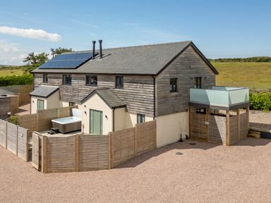 A two-story house with wooden siding and solar panels with a fenced patio and hot tub at Farmyard House in Llanfechell