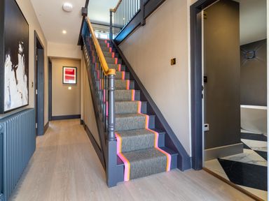A hallway with stairs covered by a runner including a wooden handrail and two paintings on the wall at Farmyard House in Llanfechell