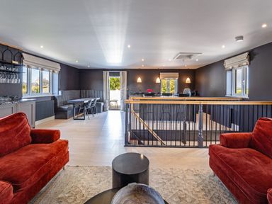 An open living and dining area with red sofas a dining table with chairs and built in seating and a kitchen island at Farmyard House in Llanfechell