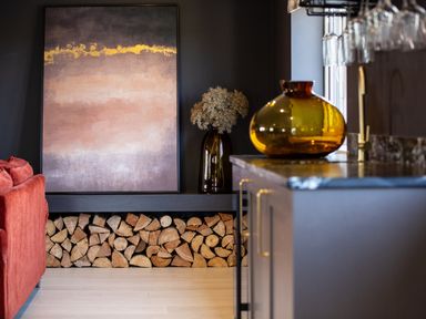 A living area with stacked firewood, an abstract painting, a dried flower arrangement in a vase, and a kitchen counter with an amber glass vase at Farmyard House Llanfechell