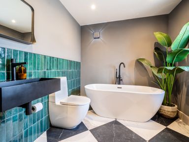 A bathroom with a freestanding bathtub toilet black sink green tiled wall and a large plant at Farmyard House Llanfechell