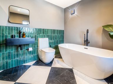 A bathroom with a white freestanding bathtub a white toilet a black wall-mounted sink with green tiled wall and a checkered floor at Farmyard House Llanfechell