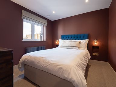 A bedroom with a bed white bedding blue headboard window with blinds and a wooden chest of drawers at Farmyard House in Llanfechell