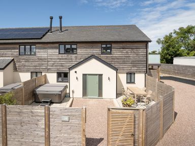 A fenced backyard with a hot tub and outdoor dining table behind a house with solar panels on the roof at Farmyard House in Llanfechell