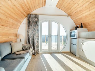 An interior room with a blue sofa a lamp a kitchen area with microwave and mini fridge and circular glass doors leading outside at Carlton Manor Pod 1 in Aldbrough