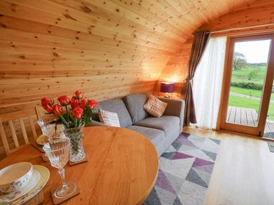 A wooden interior room with a gray sofa cushions a round wooden dining table with flowers and glassware and a door overlooking green fields at Pine Lodge in Llanddeusant near Llandovery