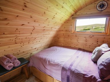 A small wooden bedroom with a bed covered in purple bedding a window a clock on the wall and three rolled towels on a bench at Pine Lodge in Llanddeusant near Llandovery