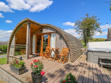 A wooden cabin with curved roof on a deck with chairs flower pots and a hot tub at Pine Lodge in Llanddeusant near Llandovery