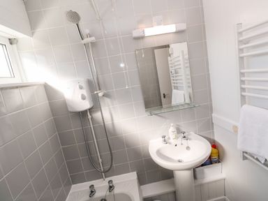 A bathroom with a shower head, electric shower unit, sink, mirror, toiletries, and towel rack at 39 Manorcombe Bungalows Honicombe Holiday Village near Drakewalls