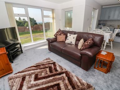 A living room with a brown leather sofa and cushions a patterned rug a TV on a stand a wooden side table and a dining area with a white table and chairs at 39 Manorcombe Bungalows in Honicombe Holiday Village near Drakewalls