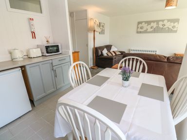 A dining area with a table set for four and a vase of flowers near a kitchen counter with appliances and a living room with brown sofas at 39 Manorcombe Bungalows in Honicombe Holiday Village near Drakewalls
