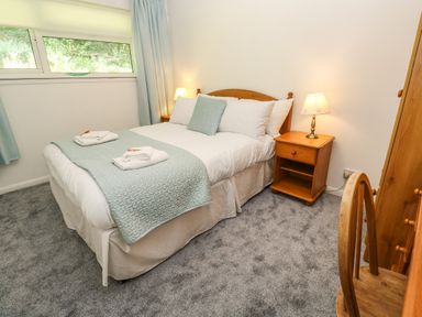 A bedroom with a double bed with white and light blue bedding two wooden bedside tables with lamps a wooden chair and wardrobe at 39 Manorcombe Bungalows Honicombe Holiday Village near Drakewalls