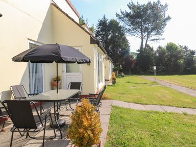 An outdoor patio with a table umbrella and four chairs next to a yellow building with a grassy area and trees at 39 Manorcombe Bungalows in Honicombe Holiday Village near Drakewalls