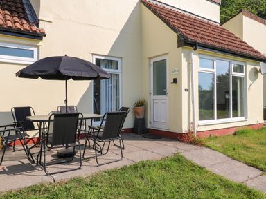 An outdoor patio with a table and four chairs under a black umbrella next to a house with a white door and large windows at 39 Manorcombe Bungalows in Honicombe Holiday Village near Drakewalls