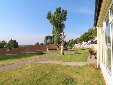 A grassy outdoor area with a paved pathway, trees, chairs, and houses at 39 Manorcombe Bungalows in Honicombe Holiday Village near Drakewalls