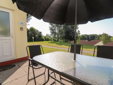 An outdoor patio area with a glass table and black chairs under a black umbrella near a yellow building at 39 Manorcombe Bungalows in Honicombe Holiday Village near Drakewalls
