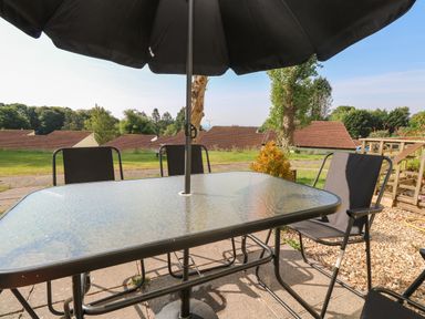 A glass outdoor table with a black umbrella and black chairs on a patio with grass and houses in the background at 39 Manorcombe Bungalows in Honicombe Holiday Village near Drakewalls
