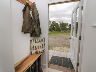 A mudroom with coats hanging on hooks a wooden bench with a cushion and boots underneath an open door showing an outdoor grassy area at Brimley Barn Kittisford near Milverton