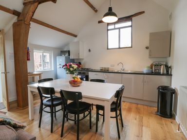 A kitchen with a white table and black chairs a fruit bowl and flowers on the table wooden beams on the ceiling and kitchen cabinets along one wall at Brimley Barn in Kittisford near Milverton