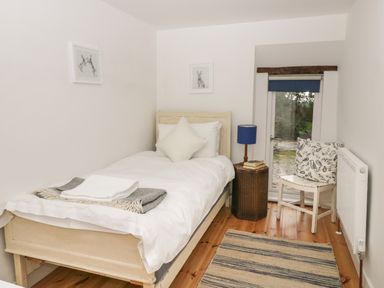 A bedroom with a single bed a small table with a lamp and books a chair with a cushion a rug on the floor and a glass door at Brimley Barn in Kittisford near Milverton