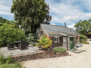 A small stone and wood house with a green door and outdoor patio furniture surrounded by trees and plants at Brimley Barn in Kittisford near Milverton