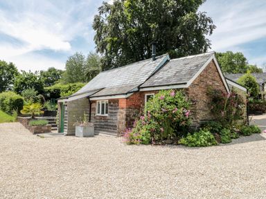 A small cottage with stone and wooden walls surrounded by plants and flowers by a gravel driveway at Brimley Barn in Kittisford near Milverton