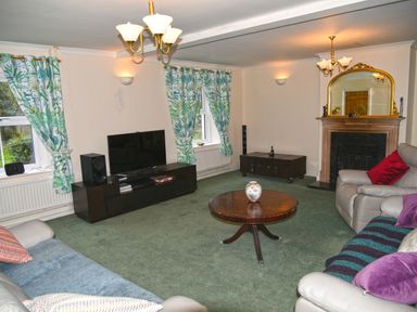 A living room with a round wooden coffee table and a television on a stand by the windows with green patterned curtains at Heathfield in Milverton