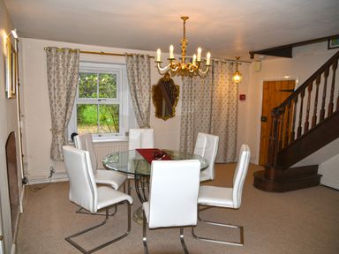A dining room with a round glass table and six white chairs near a window with curtains and a wooden staircase at Heathfield Milverton