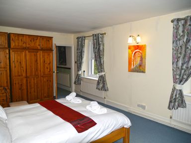 A bedroom with a bed with white linens and red runner two rolled towels wooden wardrobe and windows with patterned curtains at Heathfield in Milverton