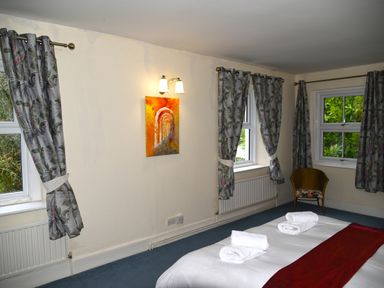 A bedroom with a bed with folded towels and a red runner two windows with curtains and a wicker chair in the corner at Heathfield in Milverton