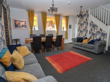 A living room with two grey sofas, yellow curtains, a wooden dining table with black chairs, a red rug, and floral wallpaper at The Old Police House in Withernsea