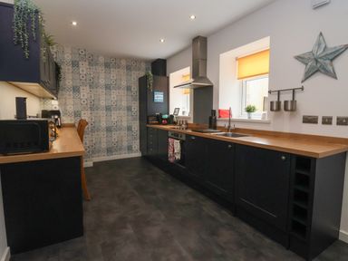 A kitchen with dark cabinets, wooden countertops, patterned backsplash, two windows with yellow blinds, and appliances at The Old Police House in Withernsea