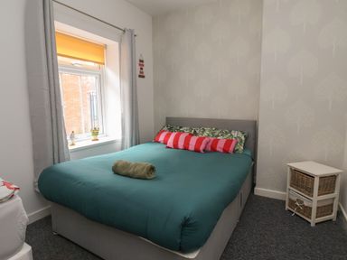 A bedroom with a double bed with patterned pillows and a rolled towel by a window with curtains and a small wicker drawer unit at The Old Police House in Withernsea