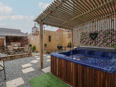 A backyard patio with a hot tub under a wooden pergola string lights small table and chairs and a barbecue grill at The Old Police House in Withernsea