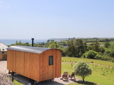 A wooden shepherds hut on wheels near two wooden chairs and a small tree with coastal landscape in the background at Little Pip in Gulval