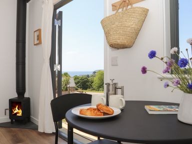 A dining area with a round black table set with pastries and mugs beside a wood burning stove and an open door showing an ocean view at Little Pip in Gulval