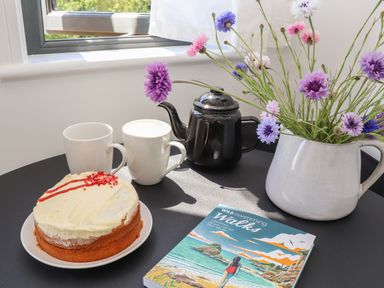A table with a cake on a plate two white mugs a black teapot a jug with flowers and a book at Little Pip in Gulval