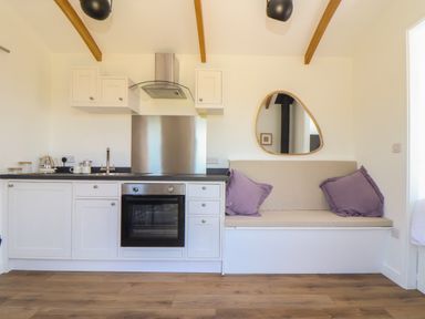 A kitchen with white cabinets oven and a beige cushioned bench with purple pillows at Little Pip in Gulval