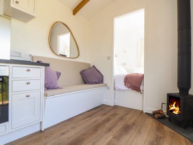 A small living area with a built-in bench with cushions a wood stove and a doorway leading to a bedroom at Little Pip in Gulval