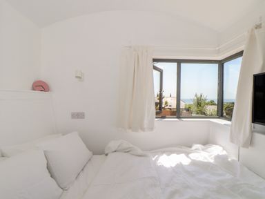 A bedroom with a white bed and pillows beside a corner window with white curtains showing outside view at Little Pip in Gulval