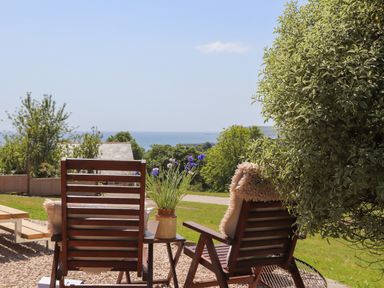 Two wooden chairs with cushions and a small table with flowers in a garden overlooking the sea at Little Pip in Gulval