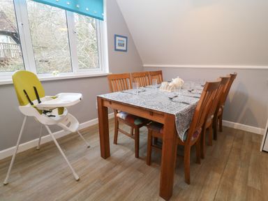 A dining room with a wooden table covered with a patterned tablecloth six wooden chairs and a green and white high chair near a window at Valley Lodge 55 Callington near Gunnislake
