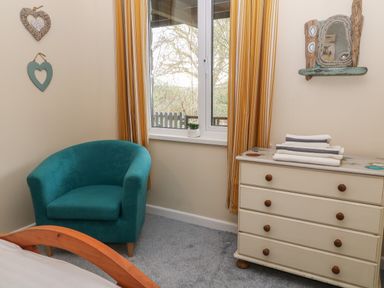 A bedroom corner with a teal armchair next to a window with yellow curtains and a white chest of drawers with folded towels at Valley Lodge 55 in Callington near Gunnislake
