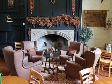 Four brown armchairs around a small round table with dried flowers in front of a large stone fireplace in a room with wooden chairs and tables Valley Lodge 55 Callington near Gunnislake